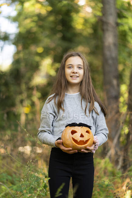 Sonriente chica sosteniendo la calabaza de Halloween mientras está de pie en el parque - foto de stock