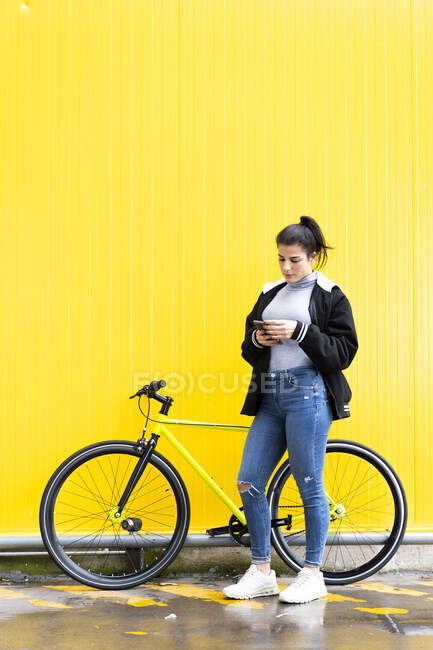 Mujer adulta utilizando el teléfono móvil mientras está de pie con la bicicleta fixie en el camino contra la pared amarilla - foto de stock