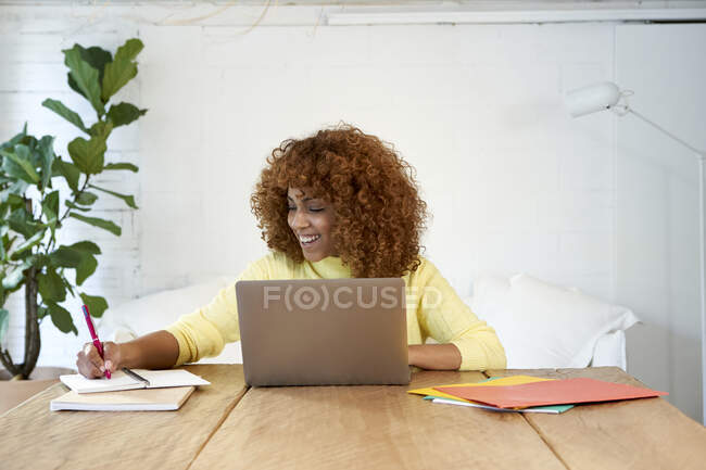 Happy businesswoman with laptop writing in notepad while working from home — Stock Photo