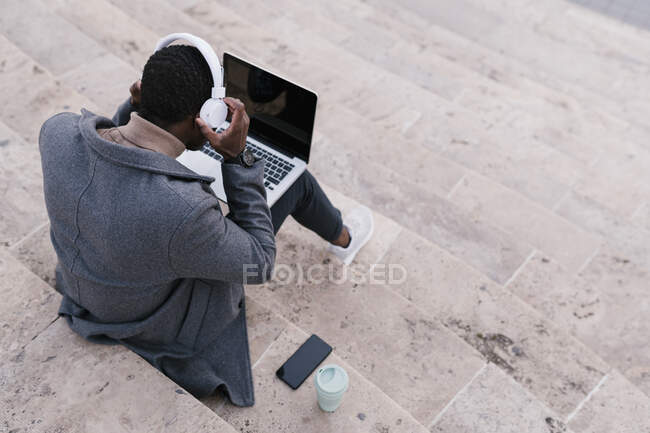 Hombre joven con auriculares usando portátil mientras está sentado en los escalones - foto de stock