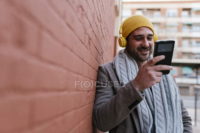 Homem com fones de ouvido sorrindo ao usar o telefone celular inclinado na parede — Fotografia de Stock