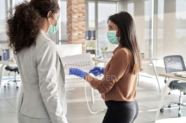 Mujer de negocios mirando a una colega mientras discute sobre los informes en el cargo durante la pandemia - foto de stock