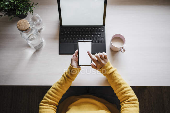 Frau benutzt Handy, während sie mit digitalem Tablet im Home Office auf dem Tisch sitzt — Stockfoto