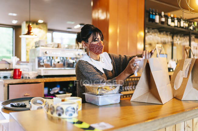 Proprietaria donna in maschera allegando ricevuta su sacchetto di carta marrone sul bancone al ristorante — Foto stock