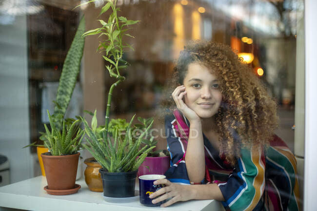 Lächelnde Frau mit Kaffeetasse an Topfpflanze gelehnt, zu Hause durch Glas gesehen — Stockfoto