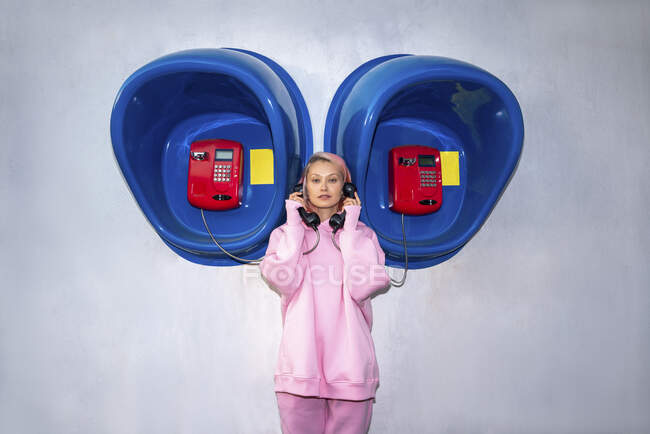 Young woman with pink hair wearing pink hooded shirt standing in front of telephone booths and holding receivers — Stock Photo