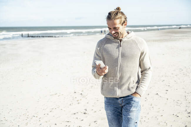Man with hand in pocket using smart phone while walking at beach - foto de stock