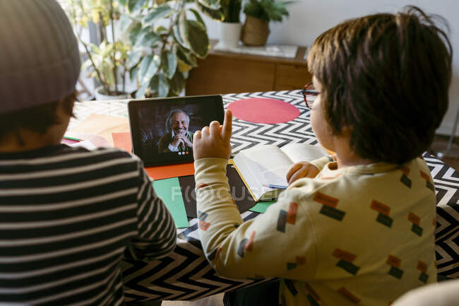 Elementary age boy pointing while e-learning through video call sitting with male friend in living room — Stock Photo