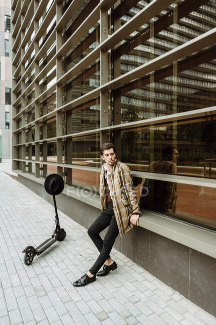 Stylish man in check jacket with push scooter sitting at modern building in city — Stock Photo