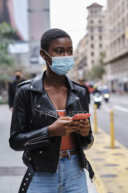 Jovem mulher usando máscara protetora usando telefone celular enquanto está na cidade — Fotografia de Stock