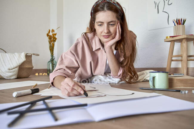Female drawing artist with hand on chin working on drawing while lying in living room — Stock Photo