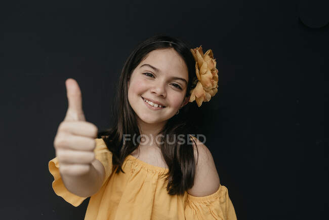 Linda chica con flor en la cabeza mostrando los pulgares hacia arriba gesto contra el fondo negro - foto de stock