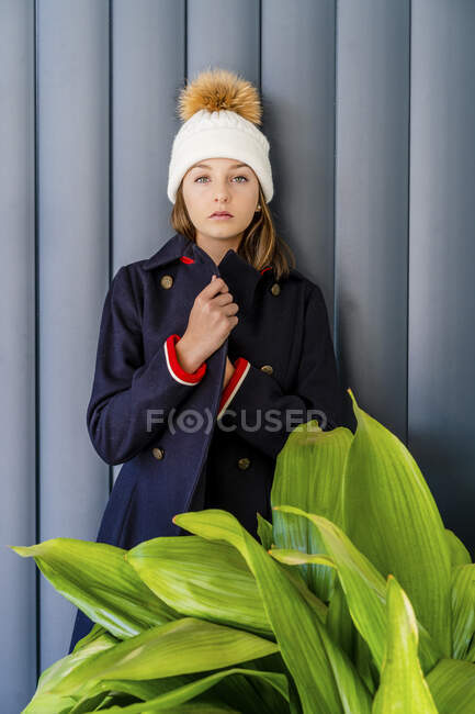 Серьёзная девочка-подросток в теплой одежде, стоящая у окна дома у стены — стоковое фото