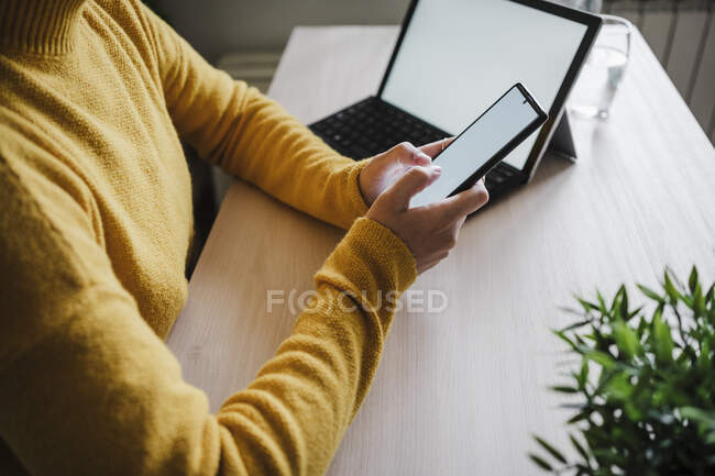 Mujer usando el teléfono móvil mientras está sentado junto a la mesa en la oficina en casa - foto de stock