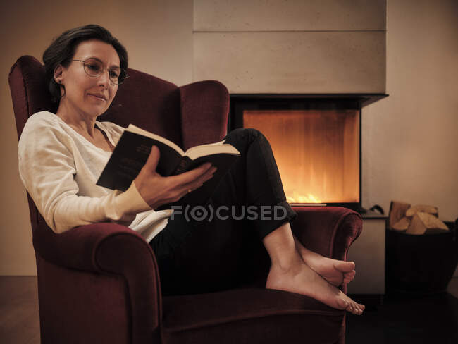 Ältere Frau liest Buch, während sie zu Hause auf einem Sessel am Kamin sitzt — Stockfoto