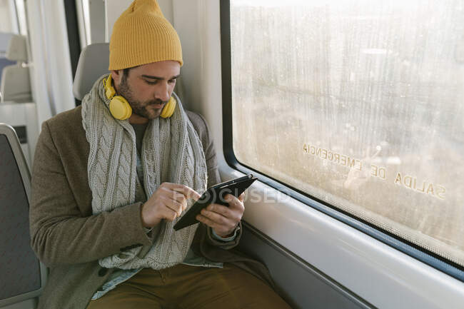 Hombre adulto medio usando sombrero de punto y auriculares usando tableta digital mientras está sentado en el tren - foto de stock