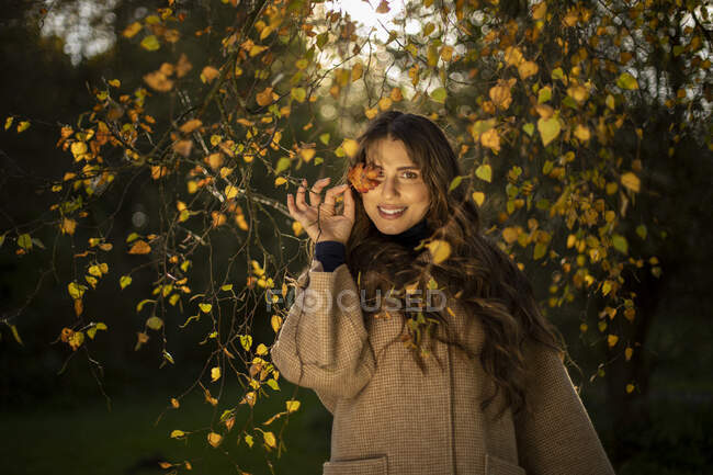 Smiling woman covering eye from autumn leaf in public park — Stock Photo