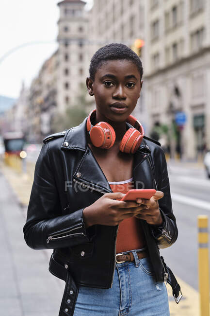 Woman wearing jacket and headphones using mobile phone while standing in city - foto de stock