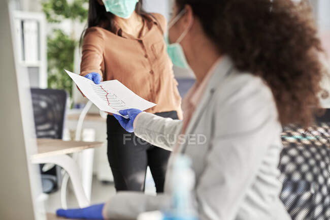 Mujer de negocios con guantes protectores entregando informes a una colega en la oficina - foto de stock