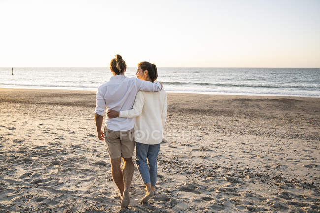 Giovane coppia con le braccia intorno a camminare in spiaggia durante il tramonto — Foto stock