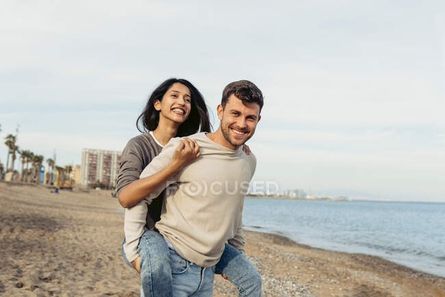Smiling boyfriend while giving piggyback ride to girlfriend at beach — Stock Photo