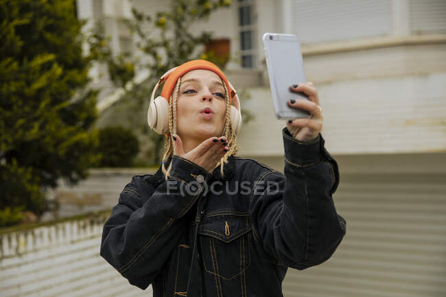 Fashionable woman blowing kiss while taking selfie through mobile phone — Stock Photo