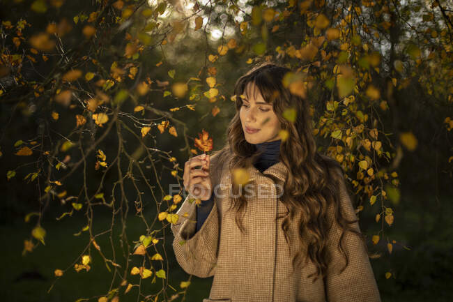 Beautiful woman wearing warm jacket looking at autumn leaf in park — Stock Photo