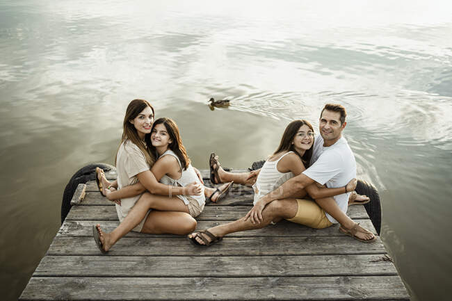 Daughters embracing mother and father while sitting on jetty against lake — Stock Photo