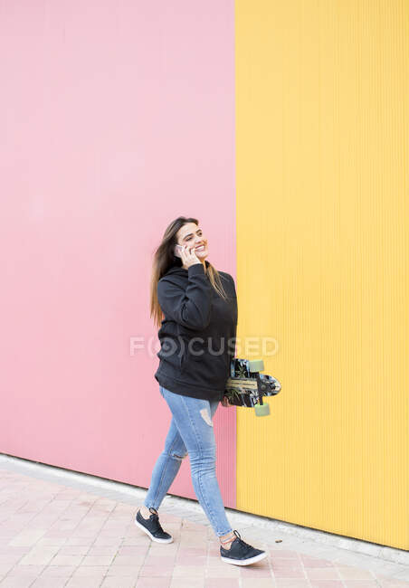 Young woman with skateboard talking on phone while walking against pink and yellow wall - foto de stock