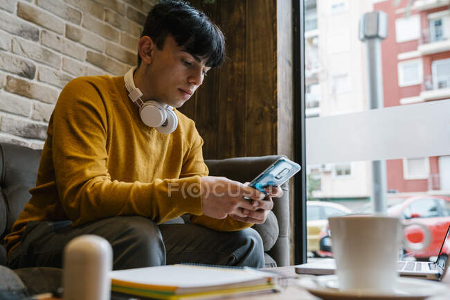 Hombre joven con auriculares usando el teléfono móvil mientras está sentado en la cafetería - foto de stock
