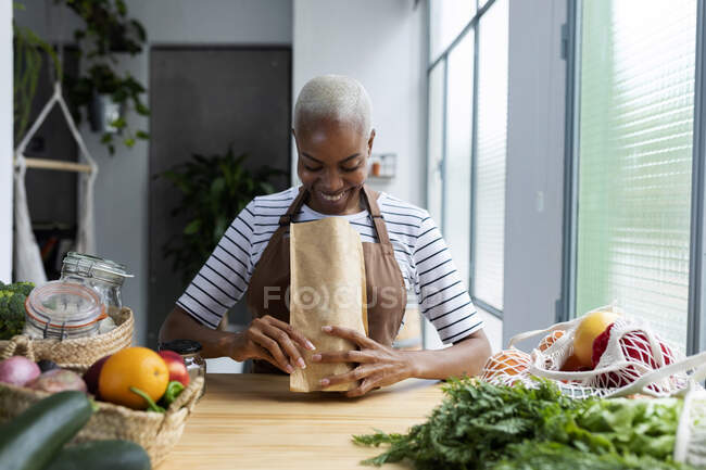 Жінка з фартухом на кухні, розпаковуючи щойно куплені органічні фрукти й овочі. — стокове фото