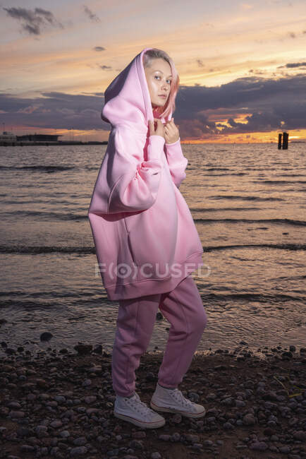 Retrato de mujer joven con camisa rosa con capucha de pie en la playa al atardecer - foto de stock