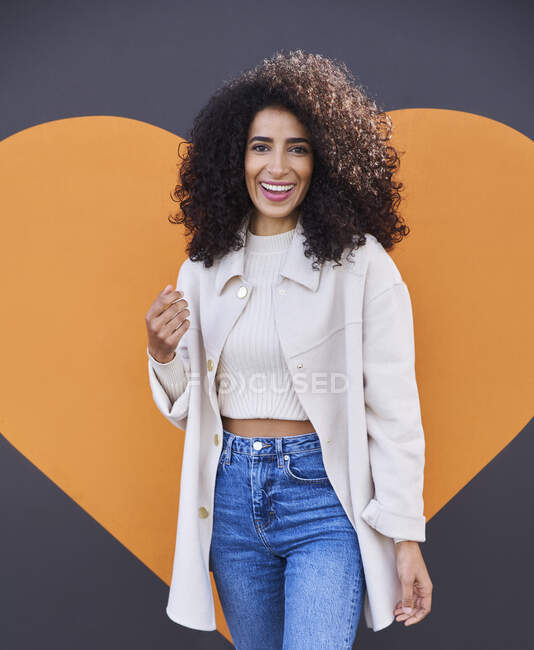 Cheerful young woman with curly hair standing against heart shape on wall - foto de stock