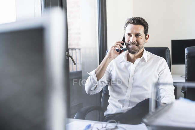 Зрілий бізнесмен розмовляє на мобільному телефоні, сидячи на стільці в офісі — стокове фото
