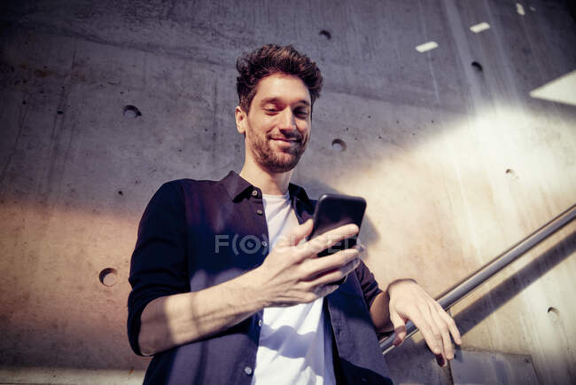 Homme d'affaires souriant utilisant un téléphone portable tout en se tenant contre le mur — Photo de stock