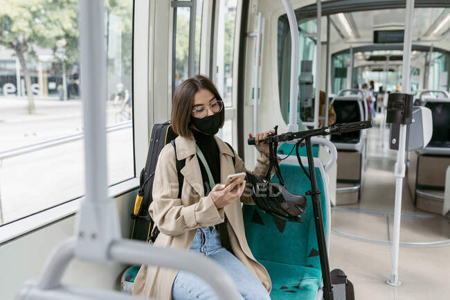 Femme adulte moyen portant un masque facial à l'aide d'un téléphone mobile tout en étant assis avec scooter électrique push dans le tramway — Photo de stock