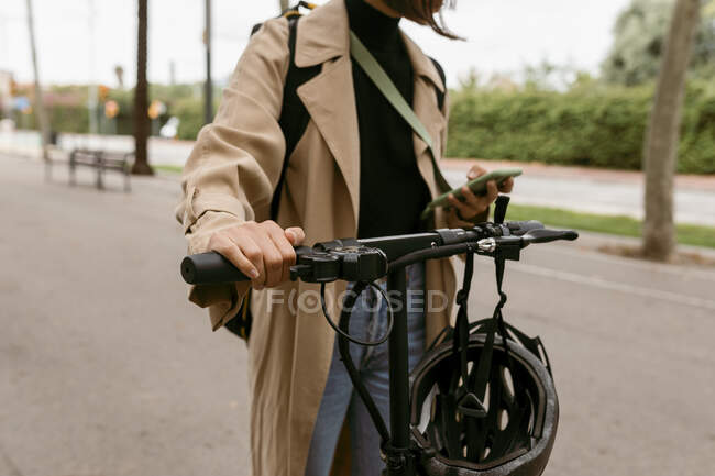 Mujer con scooter de empuje eléctrico utilizando el teléfono móvil mientras está de pie en la carretera - foto de stock
