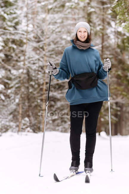 Sorrindo mulher adulta média esquiando em terra coberta de neve contra árvores na floresta — Fotografia de Stock