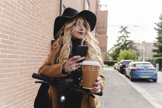Mujer joven con scooter de empuje eléctrico sosteniendo la taza de café reutilizable mientras mira hacia otro lado - foto de stock