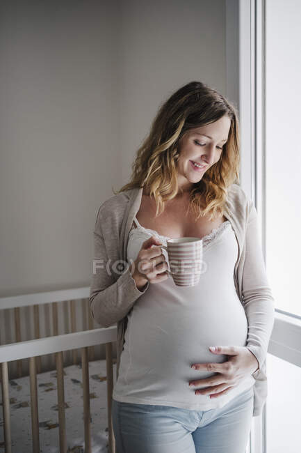 Smiling pregnant woman with hand on stomach holding coffee while standing at home — Stock Photo