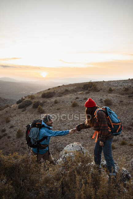 Девушка, держащаяся за руку парня, взбирающегося на скалистую гору на закате — стоковое фото