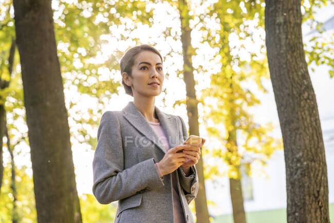 Empresaria mirando hacia otro lado mientras sostiene el teléfono inteligente de pie en el parque durante la temporada de otoño - foto de stock