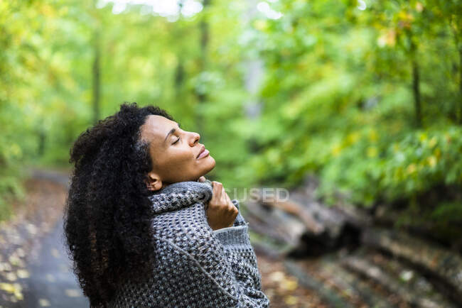 Mid adult woman wearing jacket sitting with eyes closed in forest - foto de stock
