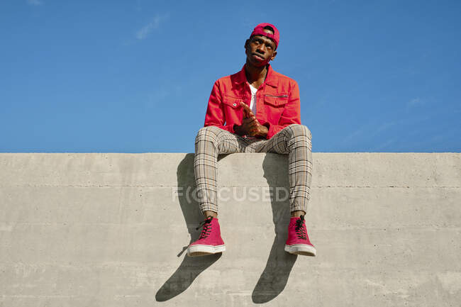 Retrato de jovem em casaco vermelho e boné de beisebol sentado na parede de concreto — Fotografia de Stock