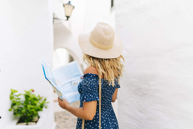 Mature woman wearing hat with map standing in Binibeca village, Minorca, Spain - foto de stock
