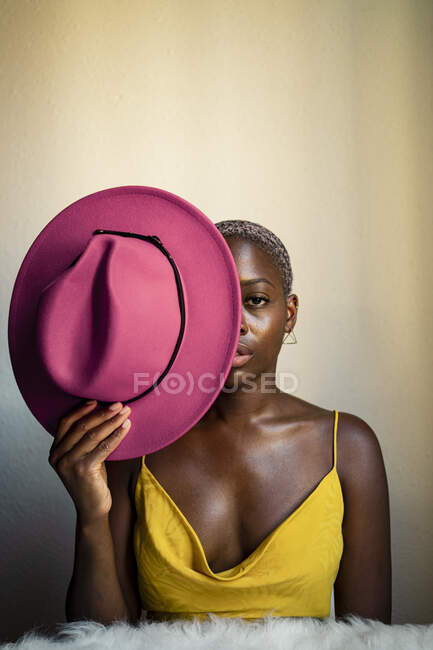 Femme confiante tenant un chapeau rose devant l'œil contre un mur blanc à la maison — Photo de stock