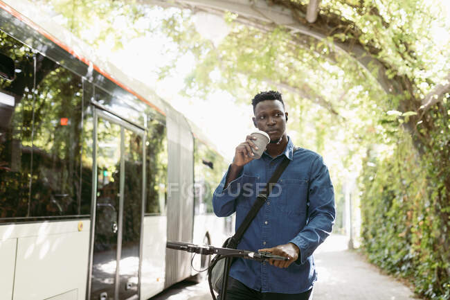Male entrepreneur with electric push scooter and reusable cup walking by bus — Stock Photo
