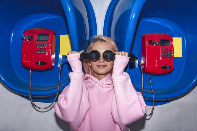Mujer joven sonriente con el pelo rosa usando camisa con capucha rosa de pie en las cabinas telefónicas, sosteniendo los receptores delante de los ojos - foto de stock