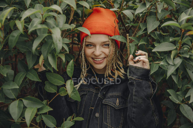 Усміхнена красива жінка в помаранчевому в'язаному капелюсі стоїть серед рослин у парку — стокове фото