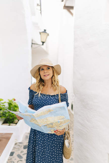 Female tourist wearing hat holding map while standing in Binibeca village, Minorca, Spain - foto de stock